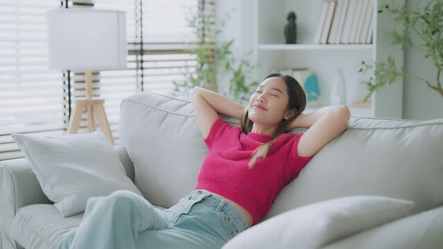 Relaxed young Asian woman enjoying rest on comfortable sofa, calm attractive girl relaxing on couch, breathing fresh air with eyes closed, meditating at home