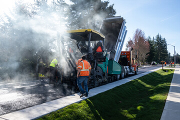 Workmen in safety clothes behind a smoking asphalt paving machine and dump truck feeding it, residential road repaving construction project
