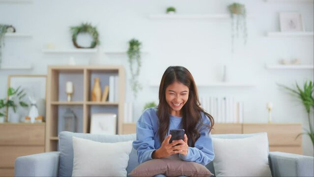 Happy young asian woman relax on comfortable couch at home texting messaging on smartphone, smiling girl use cellphone, chatting online message, shopping online from home