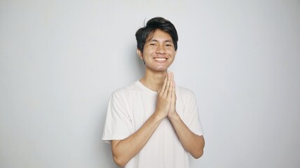 smiling young asian man in white shirt making traditional greeting gesture - obrazy, fototapety, plakaty