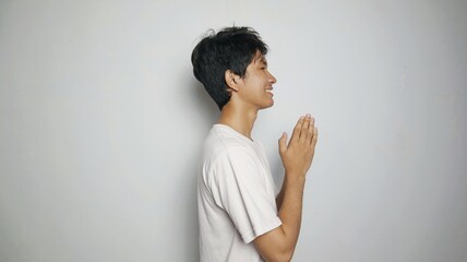 Side view of smiling young Asian man in white shirt making traditional greeting gesture on isolated...