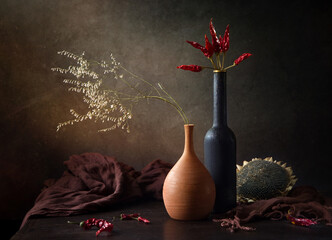 Modern still life with dry grass in a clay vase and dry hot pepper in a black bottle on a dark background