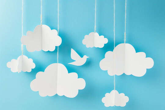 Papercraft art image of a blue background with cut-out white clouds hanging from a string, with a paper bird flying among them, Generative AI.