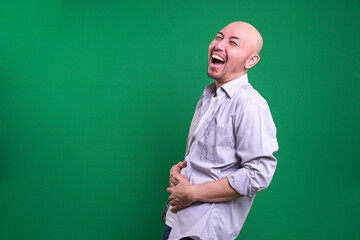 Young man in casual style standing with hands on his belly and loudly laughing isolated on green...