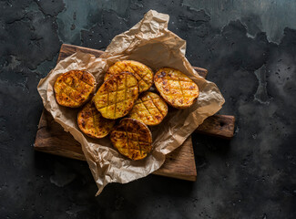 Crispy grilled potatoes on a rustic wooden chopping board on a dark background, top view
