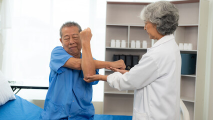  An old Asian man is doing physical therapy with the support of an elderly female nurse in the living room by a patient with a nursing attendant.