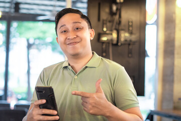 Indonesian man sitting in indoor cafe, looking at cellphone or chatting on phone with a smile, pointing at phone with a smile, wearing green polo shirt.