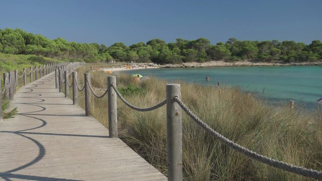 Wooden path along Son Saura sandy beach with turquoise water surrounded by green pine trees in Menorca island, Spain