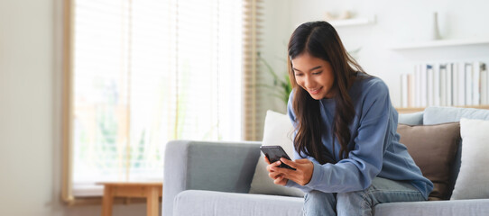 Happy young asian woman relax on comfortable couch at home, typing chat message on smartphone, smiling girl use cell phone chatting, searching information on browser wireless internet, online shopping - obrazy, fototapety, plakaty