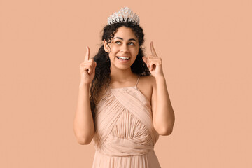 Fototapeta premium Beautiful young African-American woman in stylish beige prom dress pointing at tiara on brown background