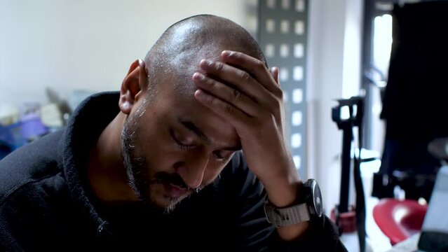 Bald South Asian British Male sits rubbing hands over head, his expression fraught with the weight of burdens, vividly capturing the palpable strain of stress and anxiety.