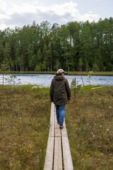 A woman walks along a narrow wooden path to a forest lake. Ecological trail in a swampy area. Traveling and hiking in forest areas. Ecological tourism and outdoor activities. Summer landscape.
