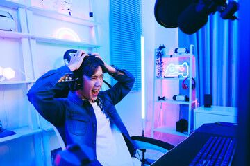 A young Asian man professional gamer wearing jeans jacket sits on a chair with hands over his head,...