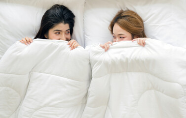 Young asian couple are peeking from under a white blanket in a bed, looking at each other with affection.