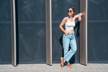 Beautiful smiling model in sunglasses. Female dressed in summer hipster white T-shirt and jeans. Posing near urban wall in the street. Funny and positive woman having fun outdoors, in sunglasses