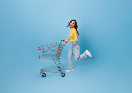 Full length happy Asian teen woman pushing empty shopping cart or shopping trolley running promotion supermarket isolated on blue background.