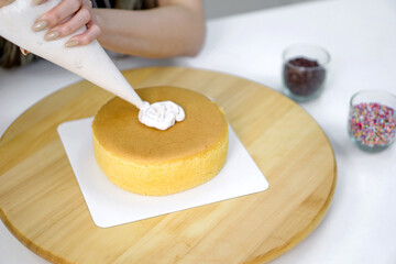 Closeup hand piping white cream on a sponge cake, creating a decorative pattern on the top of a round, light-colored cake that's placed on a wooden turntable or cake stand.