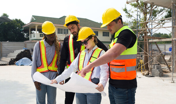 Group of construction workers gathered at a construction site reviewing some plan. Unfinished building, piles of construction material, and a partially constructed structure are in the background - Powered by Adobe