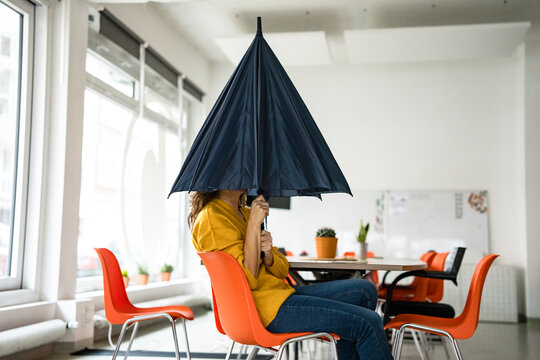 Creative businesswoman holding folded umbrella covering face at workplace