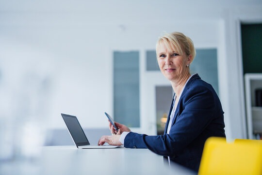 Smiling businesswoman sitting with smart phone and laptop at desk