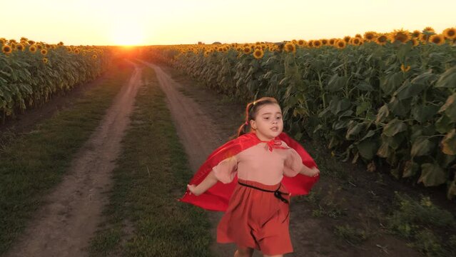 Smiling girl in red cloak superhero costume image flying at sunflower field sunset plantation. Happy female teen kid child hero running flight imagination fantasy at agriculture harvest flower meadow