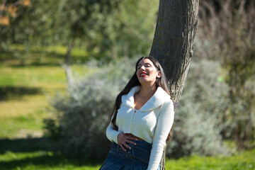 Young, beautiful Spanish brunette woman leans against the trunk of a tree in the park. The woman's lips are painted red. The girl is happy and smiling. Beauty and fashion concept.