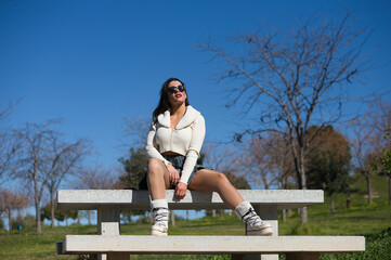 Young and beautiful Spanish brunette woman with sunglasses sitting on a park bench. The girl is casually dressed and makes different body expressions while enjoying the sunny spring day.