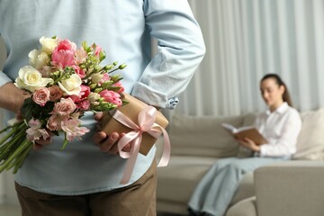 Man hiding bouquet of flowers and present for his beloved woman indoors, closeup