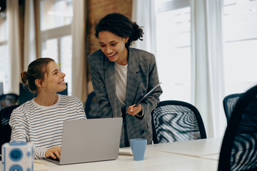 Two smiling busy female colleagues working on project together and use laptop sitting in office