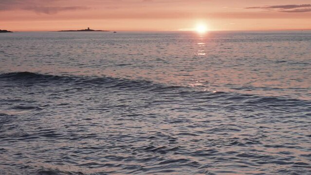 Waves lit by the warm glow of the setting sun oll slowly onto the rocky shore.