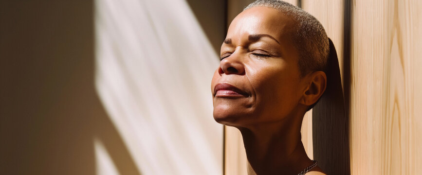 Mature bald black woman taking a deep breath and standing in morning sunlight, natural wood background