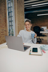 Young business woman working on laptop and drink coffee while sitting the desk in stylish office 