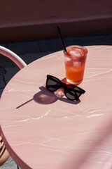 A pink round table with a glass of drink and black sunglasses on it. Minimalistc summer concept.