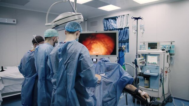 Experienced surgeons in medical gowns, caps, masks and gloves perform an operation in a modern clinic. Group of professional doctors using modern medical equipment in operating room.