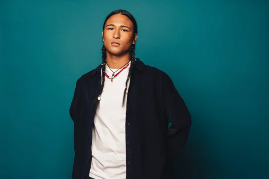 Confident native american man with stylish braided hair and jewelry standing on blue background