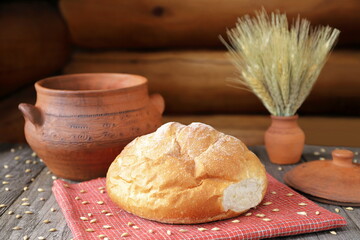 On a wooden table among the grains are a loaf of wheat bread, a clay pot and a vase with ears of wheat. 