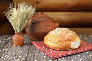 On a wooden table among the grains are a loaf of wheat bread, a clay pot and a vase with ears of wheat. 