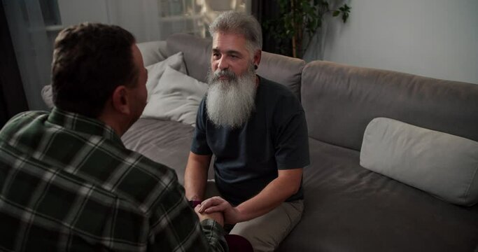 Over the shoulder A man with gray hair and a lush gray beard in a gray T-shirt communicates and holds the hand of his boyfriend a brunette with stubble in a checkered shirt sitting on a gray sofa in