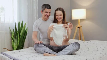 Family nesting in anticipation. Pregnant couple on bed in bedroom looking at baby clothing and talking about future child man and woman holding tiny bodysuit for their expectant kid