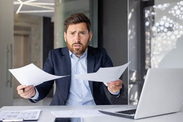Confused businessman in a suit looks angrily at paperwork in a modern office setting, feeling frustrated.