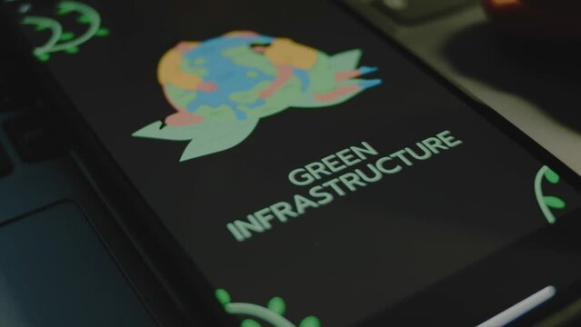 Green infrastructure inscription on black background on smartphone screen. Drawn image of two humans hugging Planet Earth. Environment concept. Male hand flapping fingers cheerfully