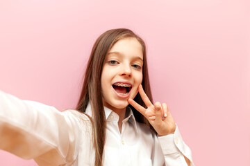 cheerful teenage girl with braces taking selfie and showing peace sign on pink isolated background,...