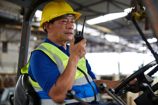 senior worker using walkie talkie and holding steering wheel on forklift truck in the factory