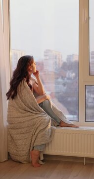 beautiful woman wrapped in a blanket sitting on the windowsill and looking outside