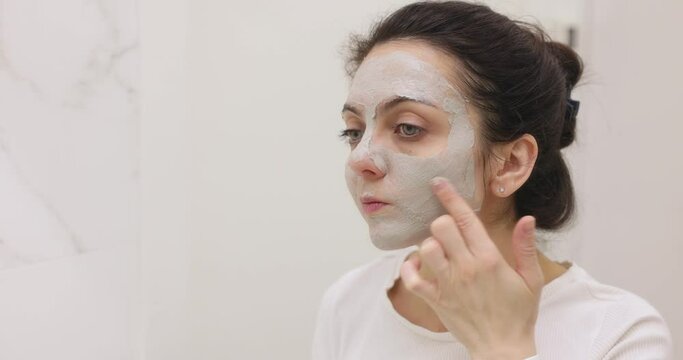 Caucasian woman looking in mirror and applying face mask in white bathroom, home spa
