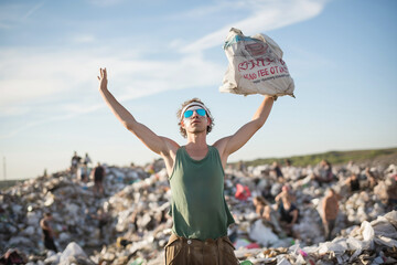 Man holding up a trash bag on a landfill site, blue sky above. - obrazy, fototapety, plakaty