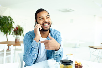 cheerful african american man with braces talking on the phone in a white cafe, a man in a denim...