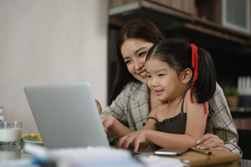Smiling Asian mother working at home with laptop sitting at kitchen table with cute daughter