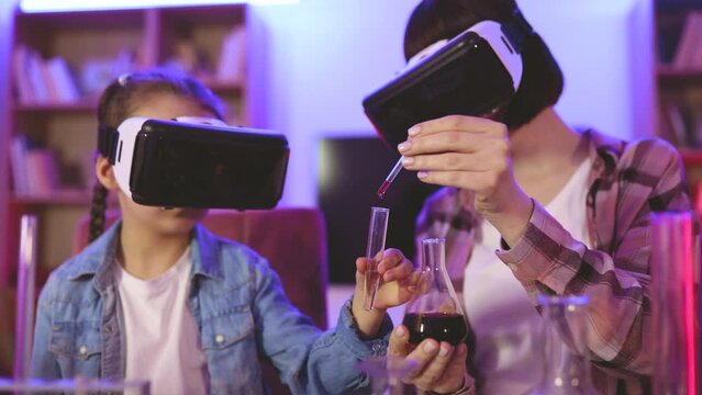 Caring young mother teaching small kid girl doing chemical experiments using vr glasses at home. Happy two generations family involved in virtual reality at evening time, children development concept.