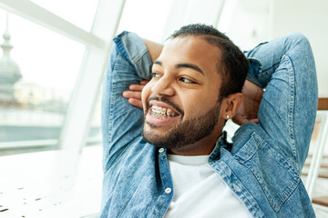 happy relaxed african american man with braces resting and looking out the window in a white room,...